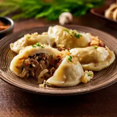 Talerz pełen pysznych pierogów z kapustą i grzybami, posypanych szczypiorkiem i skwarkami. Obok czosnek i pieprz.