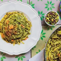 Pyszny makaron z pesto, posypany pistacjami i kawałkami łososia, prezentuje się apetycznie na białym talerzu. Obok miseczka z orzechami i listki bazylii.
