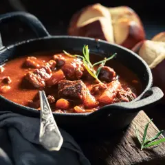 Gęsty, aromatyczny gulasz wołowy w żeliwnej patelni, z gałązką rozmarynu. Obok pieczywo. Idealny przykład, jak zagęścić gulasz.