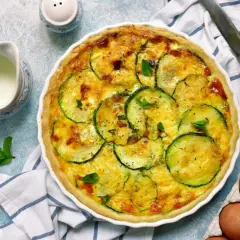 Pyszna tarta z cukinią, zapieczona ze złotym serem i świeżymi ziołami. Obok dzbanuszek mleka i jajka.