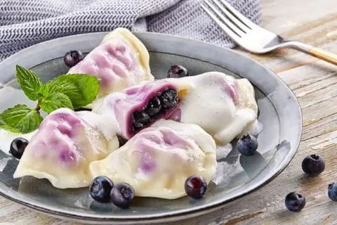 Pyszne pierogi z jagodami, polane śmietaną, ozdobione miętą i świeżymi owocami. Idealne na deser.