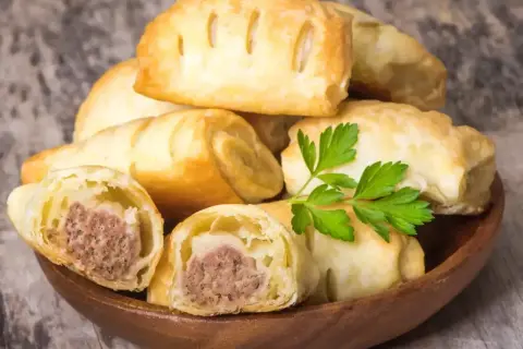 Stos pieczonego ciasta francuskiego z kiełbasą w środku, ozdobione natką pietruszki.
