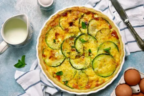 Pyszna tarta z cukinią, zapieczona ze złotym serem i świeżymi ziołami. Obok dzbanuszek mleka i jajka.