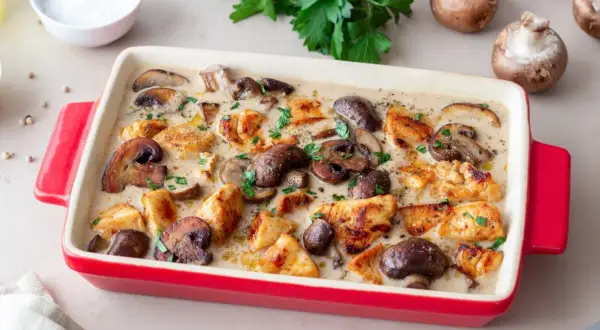 Pyszny kurczak z pieczarkami w kremowym sosie, posypany natką pietruszki, w czerwonej naczyniu żaroodpornym.