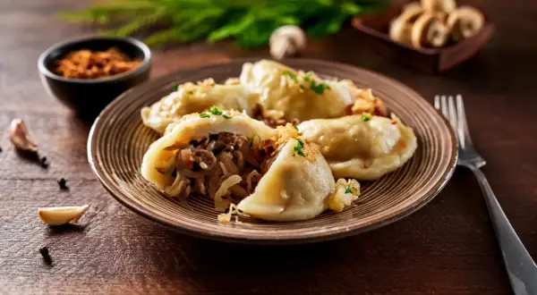 Talerz pełen pysznych pierogów z kapustą i grzybami, posypanych szczypiorkiem i skwarkami. Obok czosnek i pieprz.