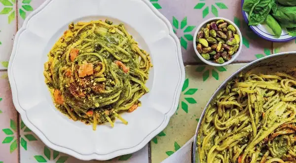 Pyszny makaron z pesto, posypany pistacjami i kawałkami łososia, prezentuje się apetycznie na białym talerzu. Obok miseczka z orzechami i listki bazylii.