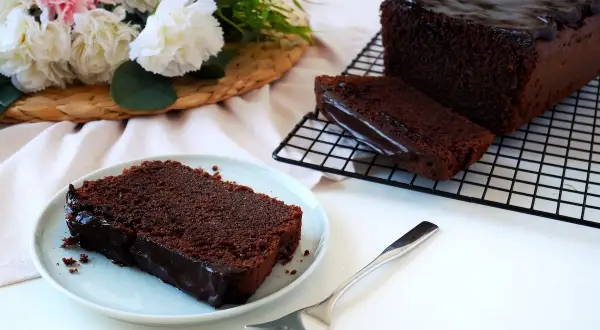 Wilgotna babka czekoladowa z polewą, podana na talerzyku z widelcem. Obok leży reszta ciasta na kratce.