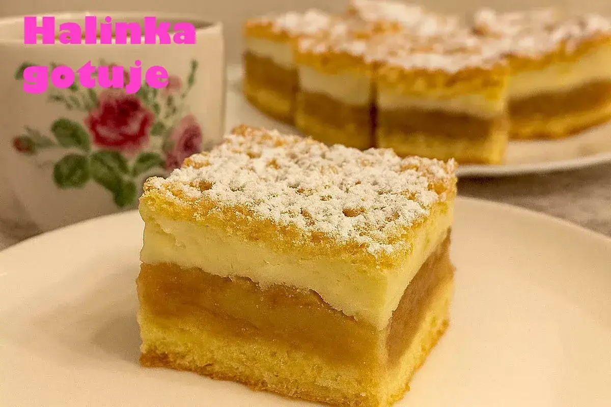 Pyszna szarlotka na dużą blachę, pokrojona w kostkę, posypana cukrem pudrem. Obok filiżanka z r&oacute;żami.