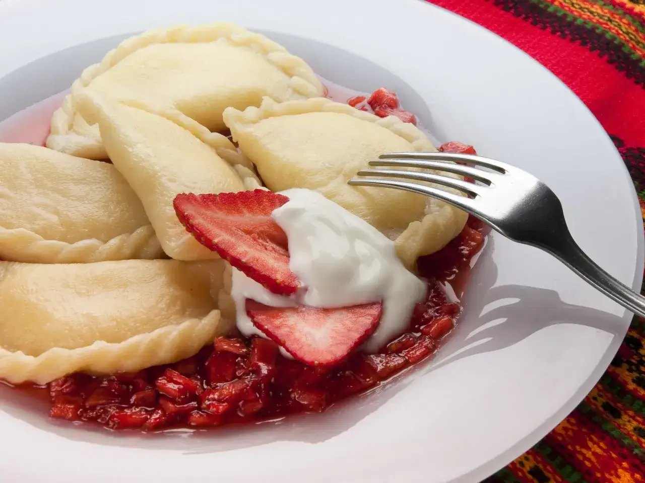 Pyszne pierogi z serem, polane sosem truskawkowym i udekorowane bitą śmietaną. Widok z widelcem na talerzu.