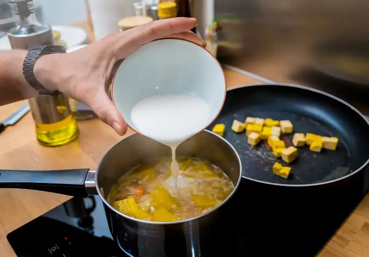 Ręka wylewa białą zawiesinę do garnka z zupą, aby ją zagęścić. Obok na patelni smażą się kostki tofu.