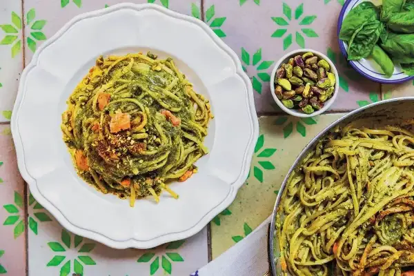 Pyszny makaron z pesto, posypany pistacjami i kawałkami łososia, prezentuje się apetycznie na białym talerzu. Obok miseczka z orzechami i listki bazylii.