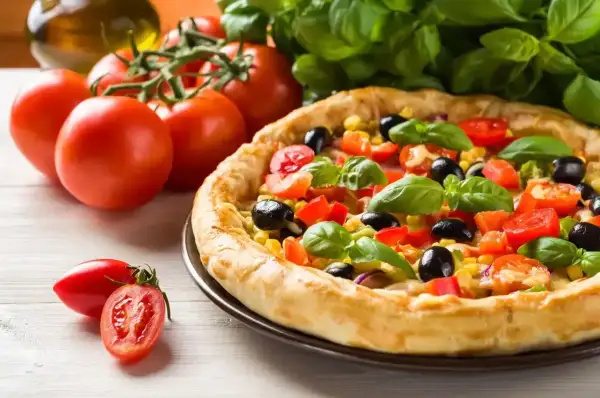 Pyszna pizza z pomidorami, oliwkami i kukurydzą, obok świeże pomidory i bazylia. Idealna na letni obiad.